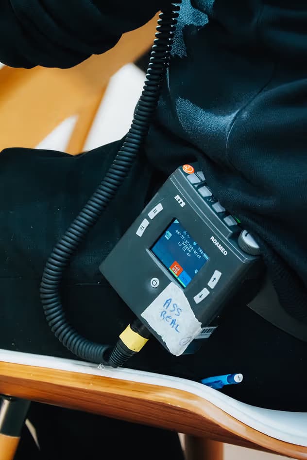 A portable electronic device attached to a person's waist with a coiled cable. The device has a display screen and buttons, labeled with a handwritten note, resting on a black garment.