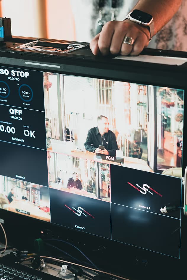 A close-up of a video editing monitor displaying multiple camera feeds and a control interface, with a person visible in one of the video windows. A hand rests on the monitor.
