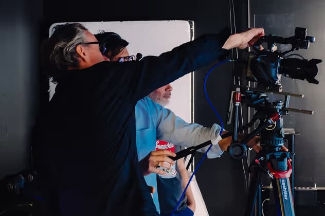 Two men working on a video production set, adjusting a camera mounted on a tripod. One man is operating the camera while the other is observing closely, both focused on their task.