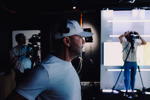A man wearing a cap is observed in profile, looking thoughtfully while standing in a studio setting. In the background, two other individuals are seen operating cameras, with bright lights illuminating a modern backdrop.