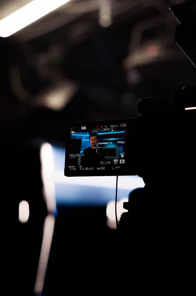 A digital camera is in focus, showing a man speaking during a broadcast. The background is blurred with a blue and black color scheme, indicating a production setting.
