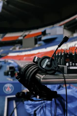 A black headset with a microphone alongside audio equipment and several coiled cables on a table, set against the backdrop of an empty sports stadium with colorful seating.