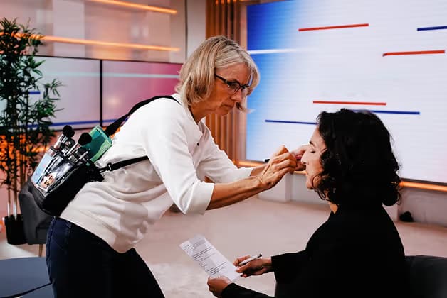 A makeup artist applying makeup to a woman in a studio setting. The artist wears a belt with makeup tools, and the woman has a clipboard in her lap as she sits in a chair. Background shows a modern studio with soft lighting.