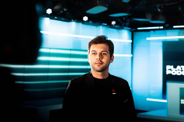 A man in a black jacket stands in a well-lit studio with a modern design. The background features blue lighting and graphics related to a podcast, creating a professional atmosphere.