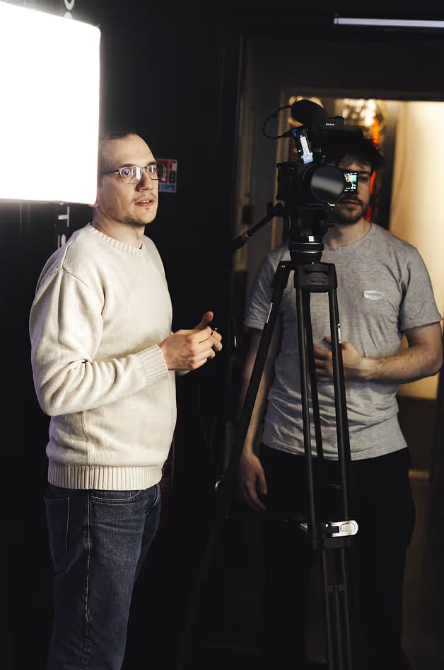 Two men in a film studio setting. One man, wearing glasses and a light sweater, is standing next to a camera on a tripod. The other man, wearing a gray t-shirt, is positioned beside him, facing the camera.