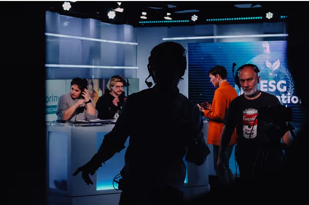 A silhouette of a crew member with a camera in the foreground, while three people are seated behind a desk in a studio setting. The backdrop displays graphics related to ESG. A man in an orange shirt is visible in the background.