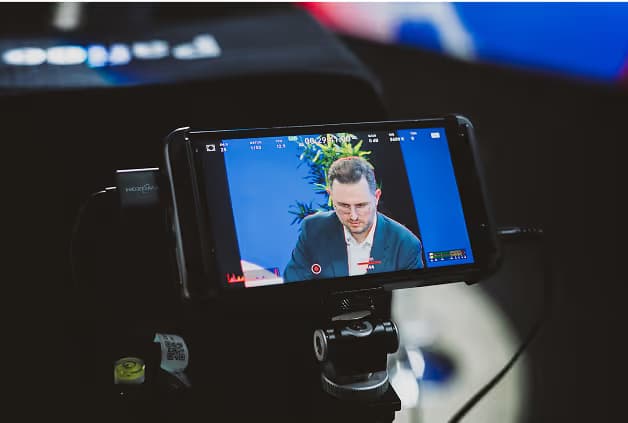 A smartphone mounted on a tripod captures a man with short hair in a blue blazer, focused while seated in front of a blue backdrop with plants. The screen shows the recording interface of the camera.