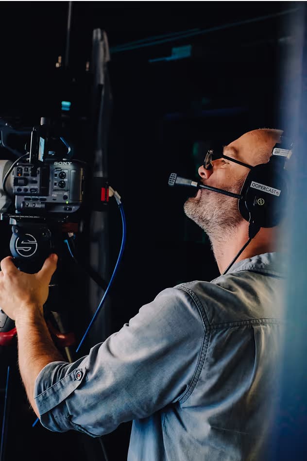 A man wearing headphones and holding a video camera while looking upwards. The setting appears to be dimly lit, emphasizing the subject in focus.