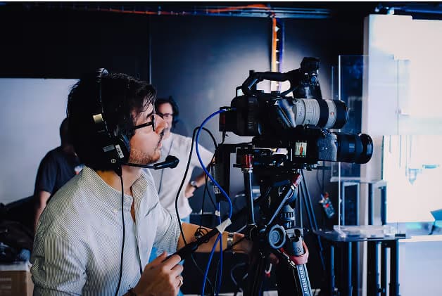 A cameraman wearing headphones operates a professional camera on a tripod. He is focused on his work in a studio environment, with additional crew members visible in the background.