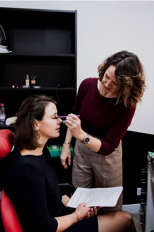 A makeup artist applying makeup to a client seated in a salon. The client is holding a paper, and the artist is using a brush while standing beside her. The background features shelves with cosmetic products.